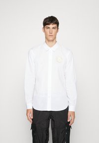 Camicia bianca a maniche lunghe in cotone, caratterizzata da un colletto button-down e una patch ricamata circolare sul lato sinistro del petto. Tessuto morbido.