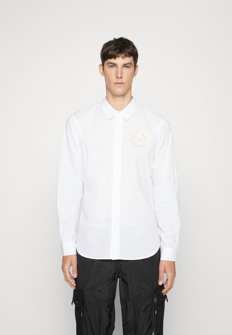 Camicia bianca a maniche lunghe in cotone, caratterizzata da un colletto button-down e una patch ricamata circolare sul lato sinistro del petto. Tessuto morbido.