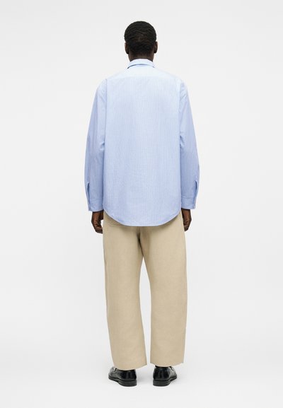 Άνδρας που κοιτάζει μακριά, φοράει πουκάμισο με γαλάζιες ρίγες και μακριά μανίκια, μπεζ παντελόνι με φαρδιά πόδια και μαύρα παπούτσια σε λευκό φόντο.