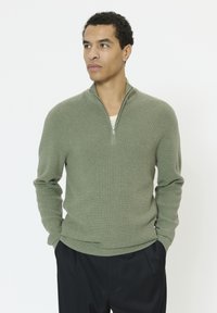 Pull-over vert en tricot avec un col zipp é, doté d'un tissu texturé et d'un ourlet côtelé, assorti à un pantalon foncé.