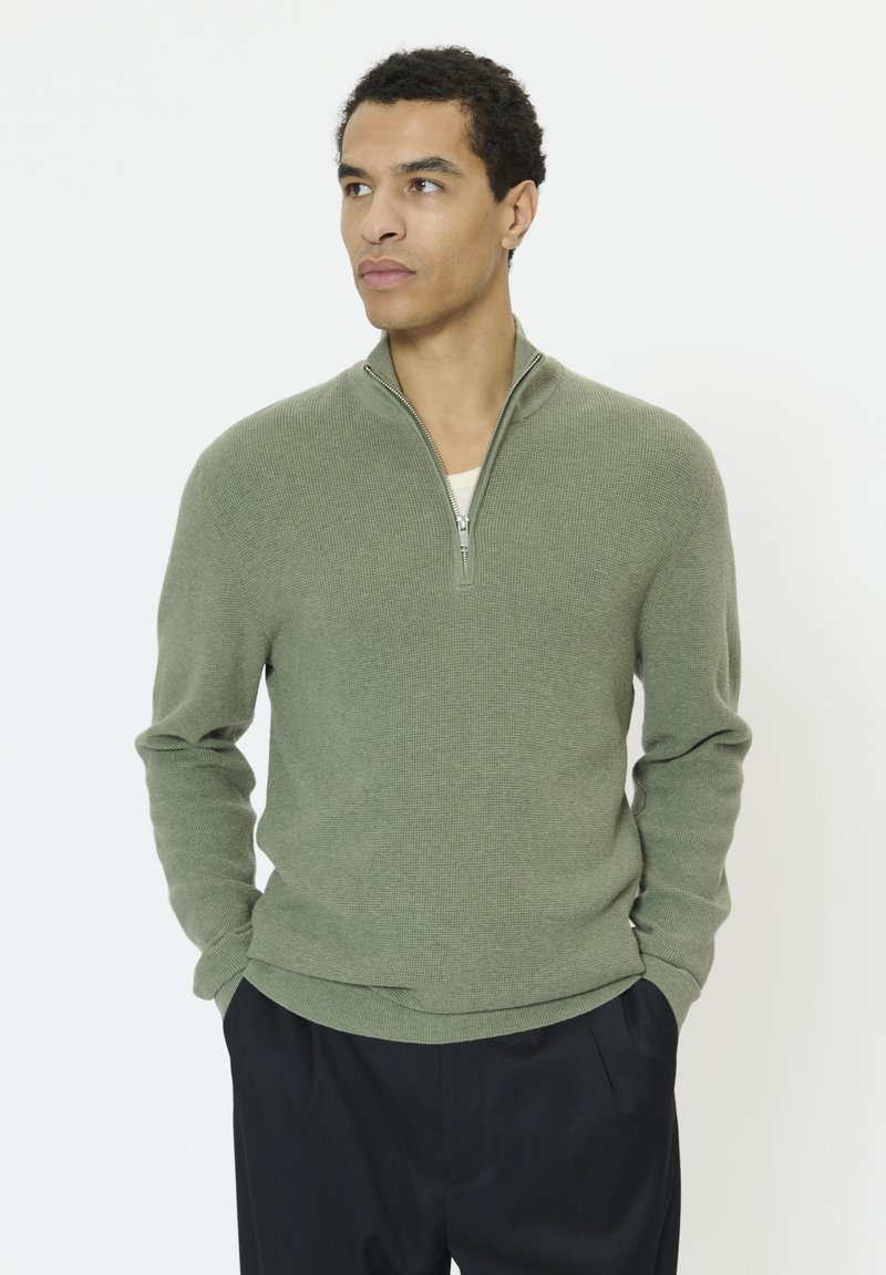 Pull-over vert en tricot avec un col zippé, doté d'un tissu texturé et d'un ourlet côtelé, assorti à un pantalon foncé.