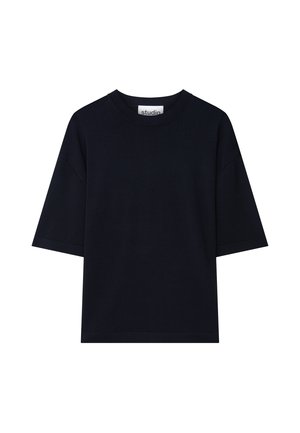 Man met kort donker haar, gekleed in een los zwart T-shirt en zwarte broek, met handen in de zakken, tegen een effen lichte achtergrond.