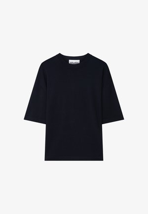Man met kort donker haar, gekleed in een los zwart T-shirt en zwarte broek, met handen in de zakken, tegen een effen lichte achtergrond.