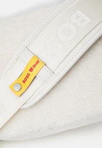 Valge pehme suss, millel on tekstureeritud kangast ülaserv, sissepressitud "BOSS" logo ning kollane silt punaste tähtedega ja metallist detail.