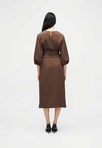 Γυναίκα με μακριά σκούρα μαλλιά φοράει καφέ midi φόρεμα με φουσκωτά μανίκια και μαύρες ψηλές γόβες, στέκεται γυρισμένη από την πλάτη σε λευκό απλό φόντο.