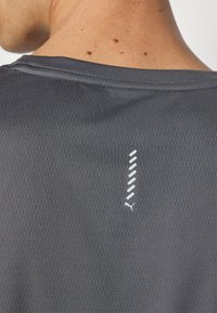 Grå atletisk t-shirt med et tekstureret, åndbart stof. Har en hvid accent med et vertikalt linjemønster og et lille logo nederst.