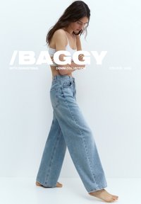 Donna che indossa jeans di denim azzurro larghi e top corto bianco, in piedi a piedi nudi contro uno sfondo bianco.