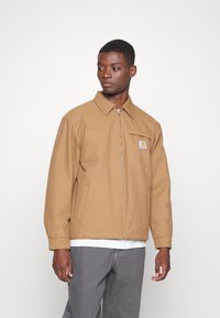 Chaqueta marrón con cremallera y diseño de cuello, que cuenta con un bolsillo de parche y un logo, fabricada con un tejido resistente. Lleva una camisa blanca debajo.