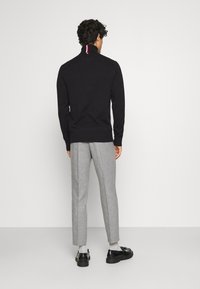 Mężczyzna w czarnej swetrze z wysokim kołnierzem z czerwonymi i białymi paskami, w szarych spodniac w kant, białych skarpetkach i czarnych skórzanych loafersach, stojący tyłem.