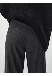 Maglione oversize nero in maglia si adagia su pantaloni grigi a righe sottili con vestibilità sartoriale e due tasche posteriori con cuciture sottili.