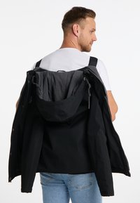 Giacca nera appoggiata sulle spalle, con un esterno liscio, cappuccio regolabile e una morbida fodera interna grigia. Jeans in denim visibili.