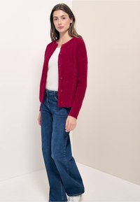 Punainen neuleliivi pyöreällä kauluksella, nappikiinnityksellä ja teksturoidulla kankaalla, yhdistettynä sinisiin leveälahkeisiin farkkuihin ja valkoisiin lenkkareihin.