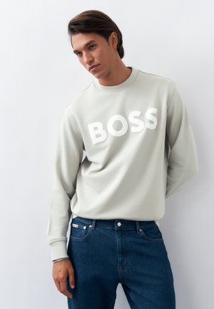 Νέος άνδρας που φοράει ανοιχτό γκρι φούτερ με τη λέξη "BOSS" σε μεγάλα λευκά γράμματα και μπλε τζιν, στέκεται μπροστά σε απλό ανοιχτό φόντο.