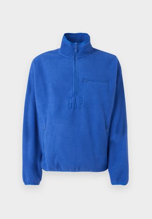 Pull polaire bleu avec fermeture éclair quart, poche zippée sur la poitrine, poches latérales zippées, col montant et logo GAP embossé au centre devant.
