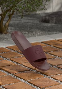 Sandale glissante bordeaux avec une large sangle arborant un logo « CK » en relief. Fabriquée en caoutchouc, elle présente une texture lisse et une semelle plate.