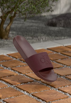 Ciabatta in colore bordeaux con una larga fascia caratterizzata da un logo "CK" impresso. Realizzata in gomma, ha una texture liscia e una suola piatta.