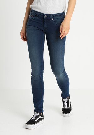 Vaqueros skinny azul oscuro con una textura suave, diseño de cinco bolsillos y cierre de botón plateado, combinados con unas zapatillas negras y blancas.