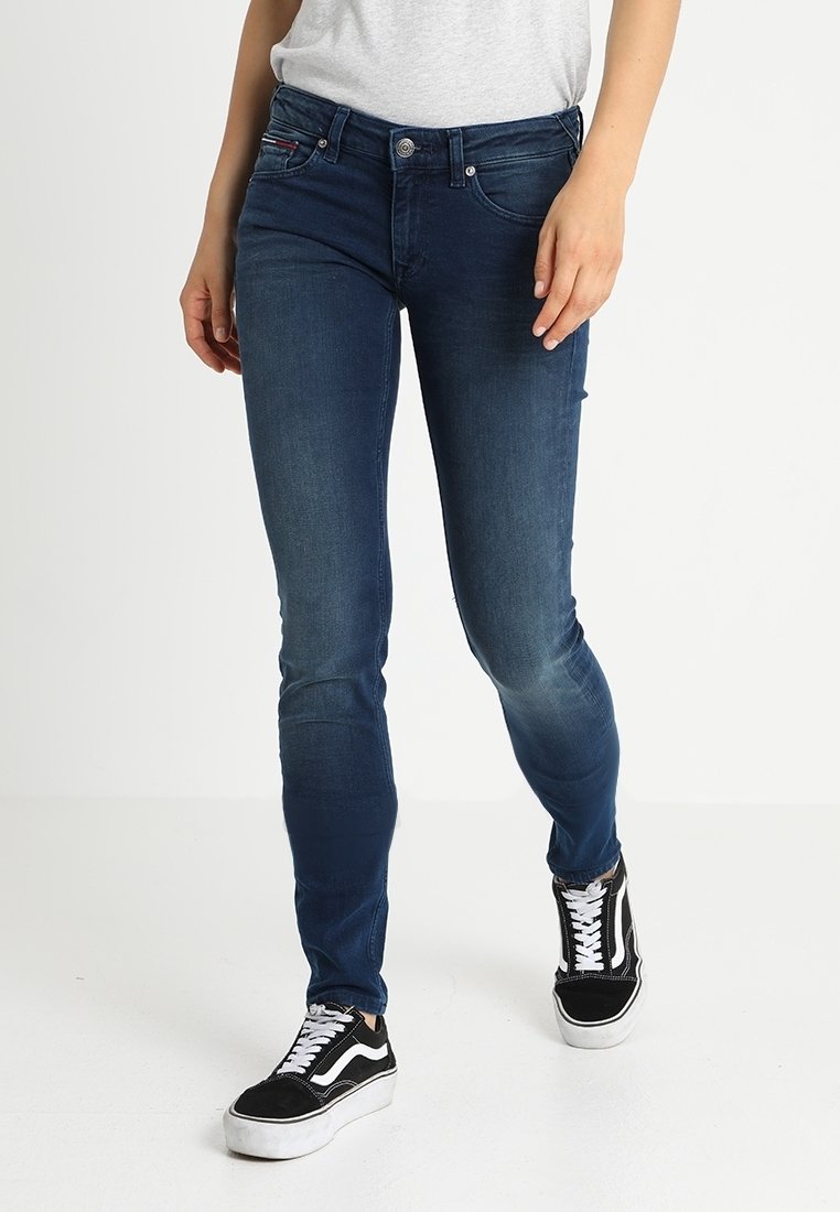 Vaqueros skinny azul oscuro con una textura suave, diseño de cinco bolsillos y cierre de botón plateado, combinados con unas zapatillas negras y blancas.