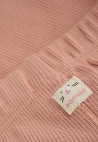 Close-up van roze ribbelstof met een klein wit geweven label met een kattengezicht en de tekst "NORMES."