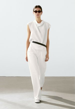 White sleeveless top with draped neckline, paired with high-waisted white trousers and a black belt. Model wears dark sunglasses.