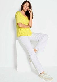 Gelbes, drapiertes Oberteil aus leichtem Stoff, kombiniert mit weißen, knöchellangen Hosen und hellen Sneakers. Modell sitzt auf einem weißen Podest.