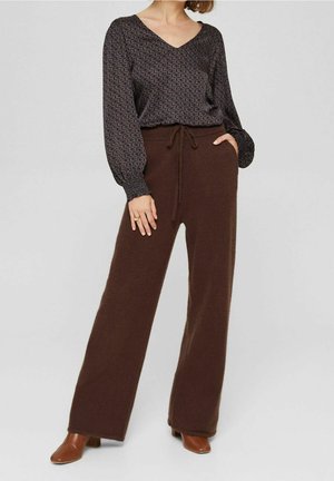 Kobieta nosząca wzorzystą bluzkę z długim rękawem włożoną do brązowych, szerokich spodni z wysokim stanem z sznurkiem i brązowych botków za kostkę.