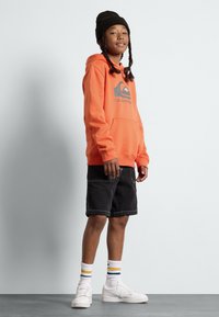 Orange hoodie med en logotyp, svarta shorts, vita sneakers och randiga strumpor. Hoodien har en framficka och en avslappnad passform.