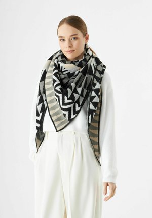 Vrouw in witte trui en broek met een grote sjaal in een zwart, grijs en beige geometrisch patroon om haar nek gedrapeerd.