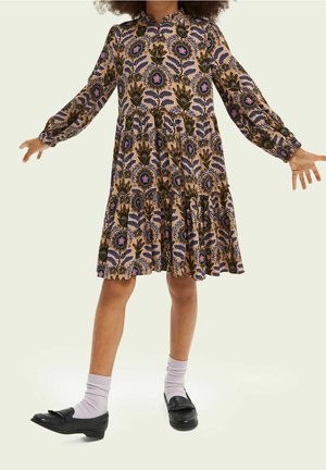 Brun blomsterkjole med lange ærmer, samlet kant og krave. Sorte loafers parret med lys grå sokker. Baggrunden er lysegrøn.