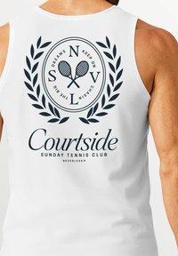 Weißes ärmelloses Shirt mit einem runden Grafikdesign, das Tennisschläger, Lorbeerblätter und den Text "Courtside Sunday Tennis Club" in Marineblau zeigt.