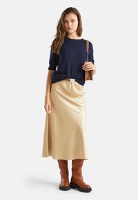 Granatowy sweter krótki z rękawami, beżowa satynowa spódnica maxi, brązowe kozaki sięgające kolan z czarnymi podeszwami oraz mała brązowa torebka na łańcuszku.