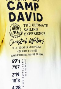 Camiseta amarilla con texto negro, que incluye "CAMP DAVID" y detalles de temática náutica; incluye coordenadas y frases sobre navegación.