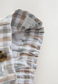 Detail béžovej a bielej károvanej látky s elastickým pásom a bielym nastaviteľným gombíkom vo vnútri.