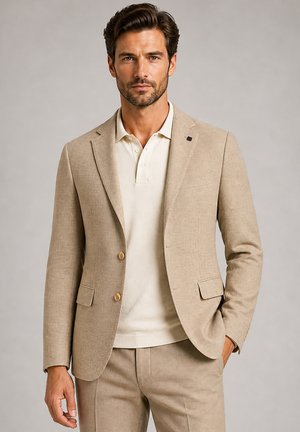 Man wearing beige suit jacket and matching trousers over cream polo shirt standing with one hand in pocket, neutral background.