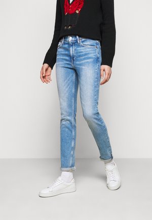 Persoon draagt lichtblauwe slim-fit jeans, witte sneakers en een zwarte trui met een rode afbeelding, staand op een lichtgrijze vloer.