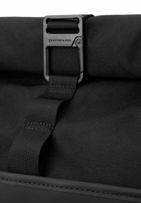 Černá textilní taška s matnou černou kovovou sponou a nastavitelným popruhem. Povrch má strukturovaný povrch s odolným šitím.