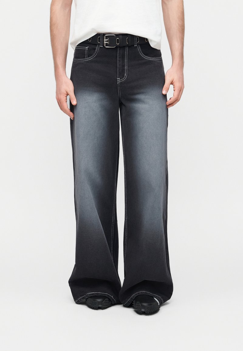 Άνδρας με μαύρο τζιν παντελόνι φαρδιάς γραμμής με ξεθωριασμένο γκρι μπροστά, μαύρη ζώνη, λευκή μπλούζα, στέκεται μπροστά σε απλό φωτεινό φόντο.