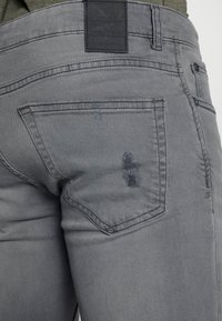Jeans de denim gris con una textura desgastada, decoloración sutil y diseño de cinco bolsillos. Etiqueta de cuero negra en la cinturilla.