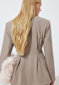 Blazer beige a medida con hombreras estructuradas, espalda plisada y detalle de suave pelo sintético rosa. El cabello cae en ondas sueltas sobre el hombro.