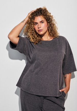 Kroeshaar vrouw die een losse donkergrijze korte mouw top en bijpassende broek draagt, poseert met één hand in haar zak en de andere raakt haar hoofd aan.