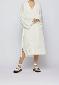 Abito bianco a maniche lunghe con scollatura a V e maniche larghe, caratterizzato da un motivo a maglia testurizzato e fessure laterali. Abbinato a sandali bianchi chunky.