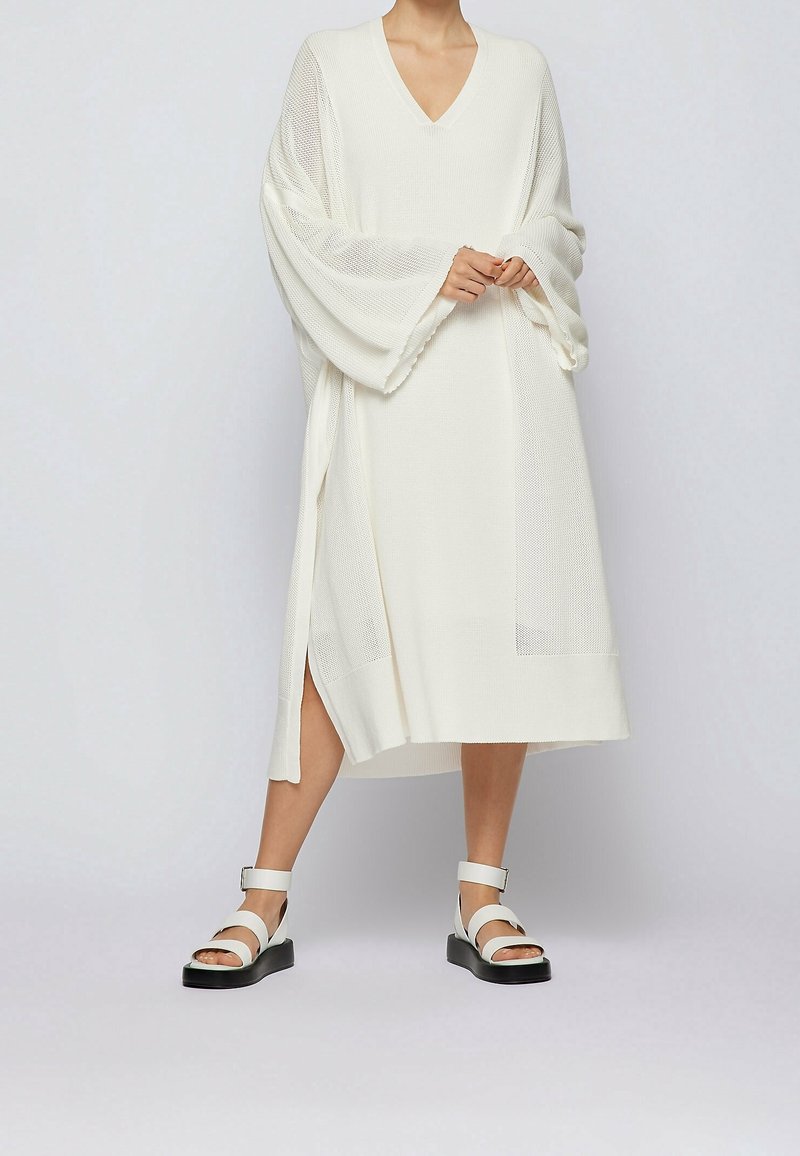 Abito bianco a maniche lunghe con scollatura a V e maniche larghe, caratterizzato da un motivo a maglia testurizzato e fessure laterali. Abbinato a sandali bianchi chunky.