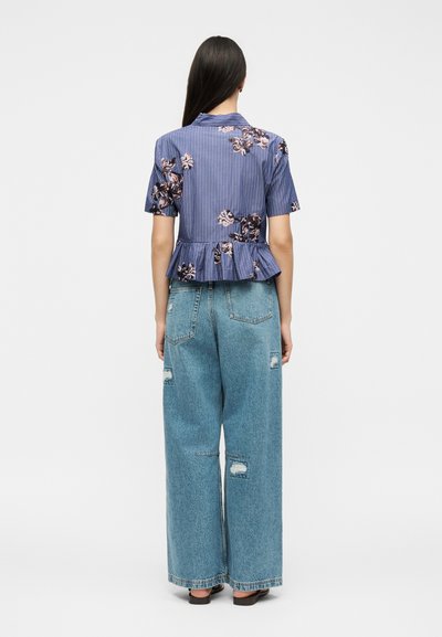 Femme aux longs cheveux foncés, portant un chemisier bleu à manches courtes à fleurs et un jean large usé, debout dos à la caméra.