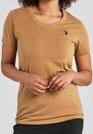 Žena na sobě má dobře padnoucí světle hnědé tričko s krátkým rukávem, na levé straně hrudi je malé vyšívané černé logo.