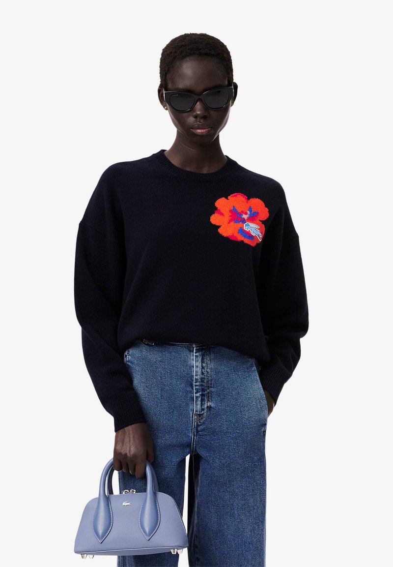 Marineblauer Strickpullover mit einem großen roten Blumenmuster, kombiniert mit hellblauen hoch geschnittenen Jeans und einer kleinen strukturierten Handtasche.