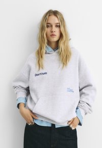 Felpa grigia con cappuccio con la scritta "Don't hate" ricamata in blu navy, indossata sopra una camicia a quadri blu chiaro. Jeans scuri e anelli visibili.