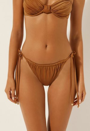 Femme portant un bikini marron froncé avec des bas à nouer sur les côtés, visible de la poitrine jusqu'aux cuisses, sur un fond uni.