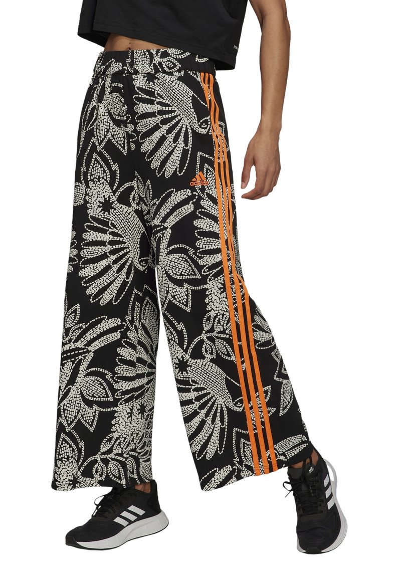 Pantalon noir à jambes larges avec motifs floraux blancs et trois bandes latérales orange, porté avec des baskets noires et blanches et un top court noir.