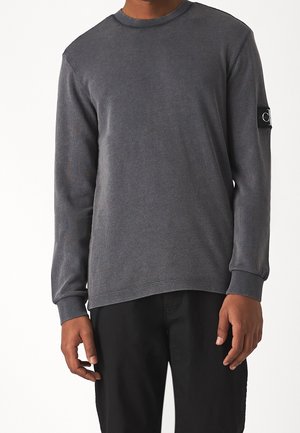 Homme portant un pull à manches longues gris foncé texturé avec un patch de marque sur le bras supérieur gauche et un pantalon noir.