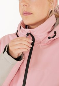 Rosa Winterjacke mit hohem Kragen, schwarzem Reißverschluss und verstellbaren Kordeln an der Kapuze. Strukturierter Stoff und kontrastierende graue Bündchen.